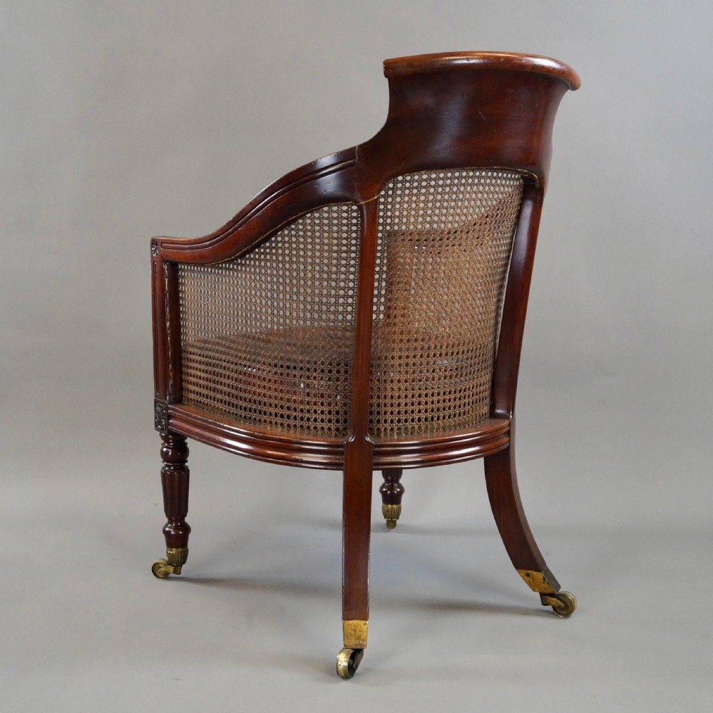 A Regency mahogany bergere armchair with a carved cresting rail, woven cane back and sides, a buttoned brown leather seat cushion, and brass castors. View VII
