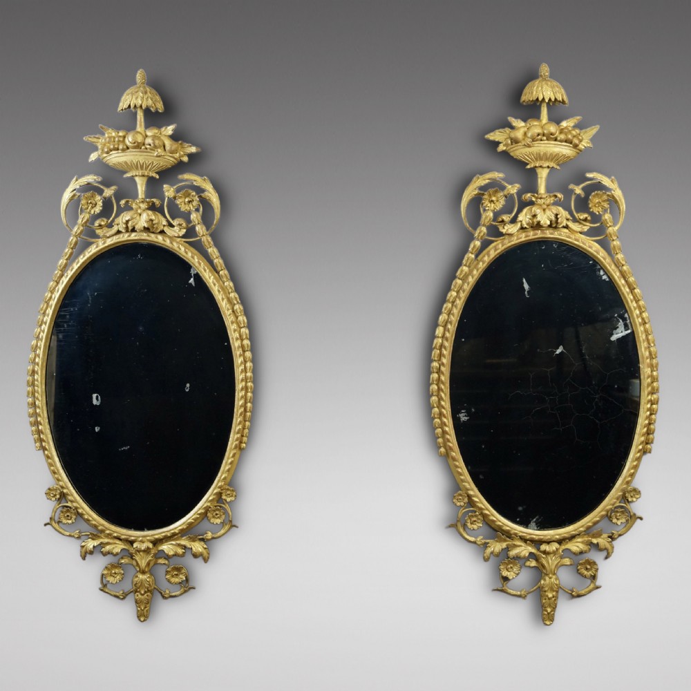 A pair of 18th-century oval giltwood wall mirrors featuring carved floral surrounds and fruit-filled urn finials.