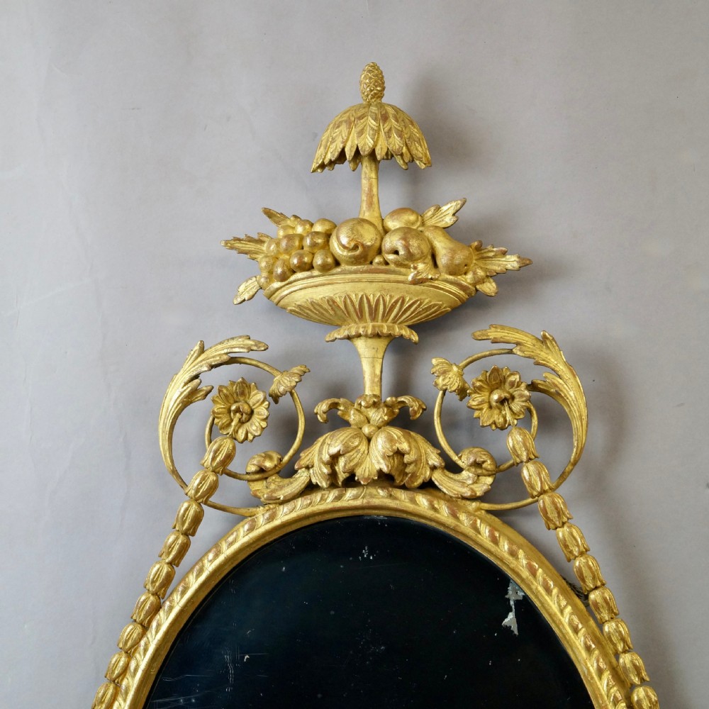 A pair of 18th-century oval giltwood wall mirrors featuring carved floral surrounds and fruit-filled urn finials. View II