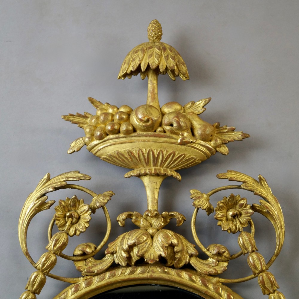 A pair of 18th-century oval giltwood wall mirrors featuring carved floral surrounds and fruit-filled urn finials. View V