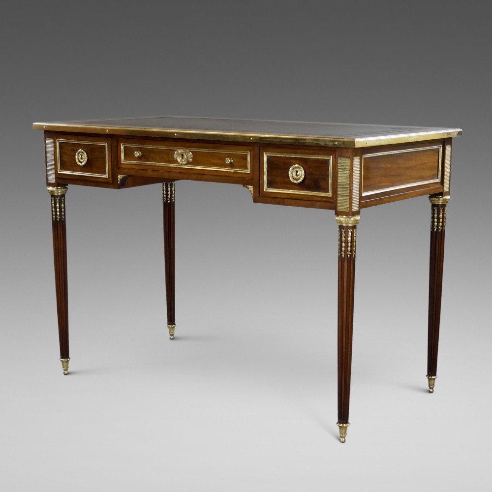 A mahogany writing desk featuring three frieze drawers with brass mounts, a leather-inlaid top, and four tapered fluted legs tipped with brass feet.