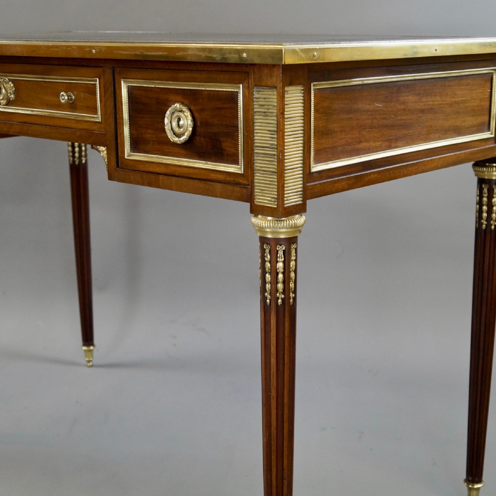 A mahogany writing desk featuring three frieze drawers with brass mounts, a leather-inlaid top, and four tapered fluted legs tipped with brass feet. View V