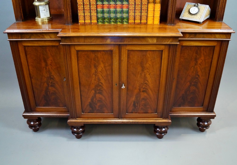 A mahogany breakfront bookcase featuring open upper shelving, flame mahogany panelled lower cupboard doors, and turned feet. View III