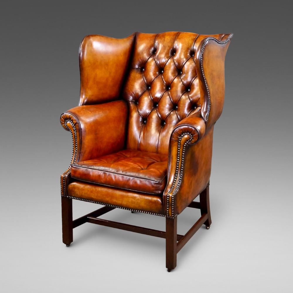 A George III period tan leather wingback armchair featuring deep button tufting, brass stud detailing, scrolled arms, and square mahogany legs united by stretchers.