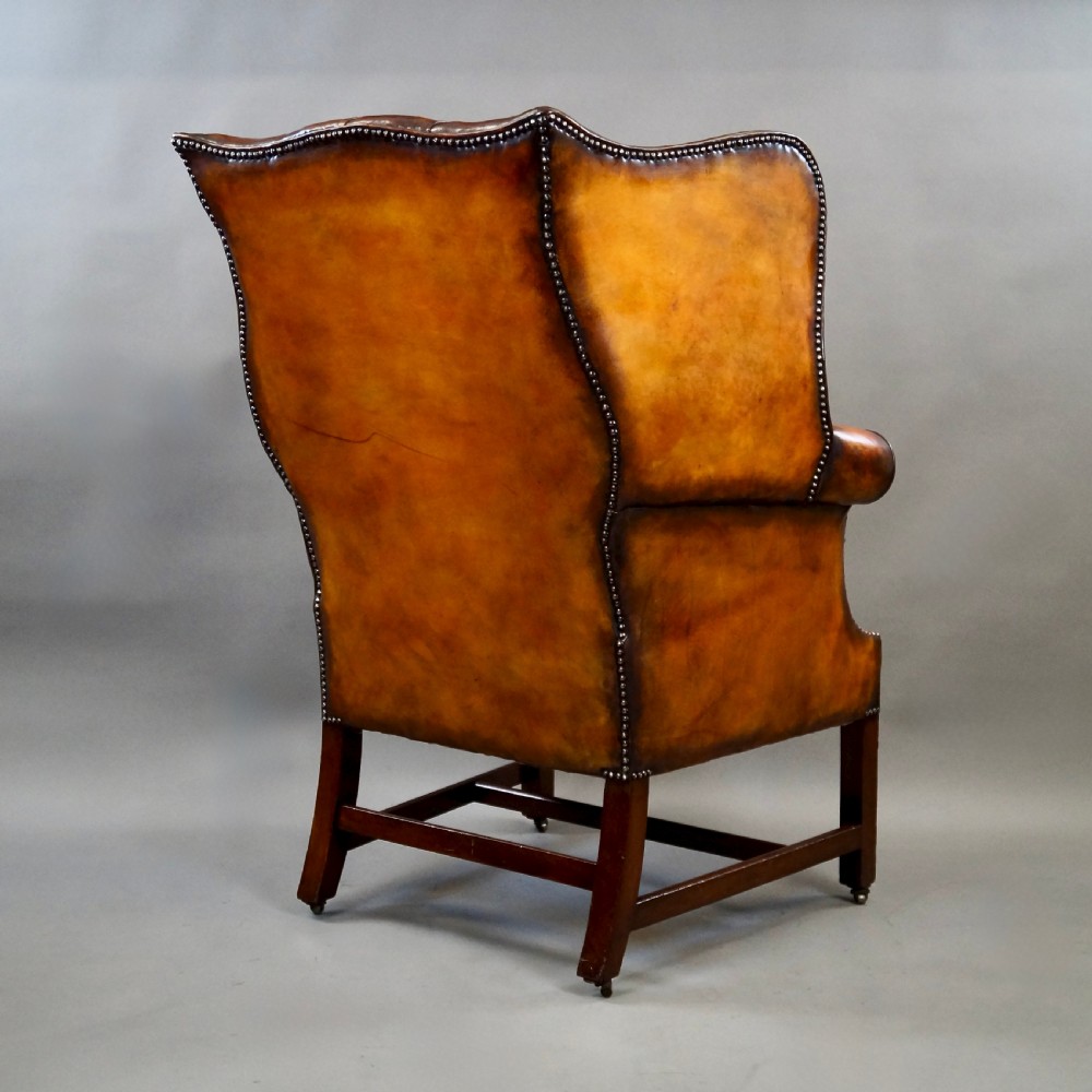 A George III period tan leather wingback armchair featuring deep button tufting, brass stud detailing, scrolled arms, and square mahogany legs united by stretchers. View IV