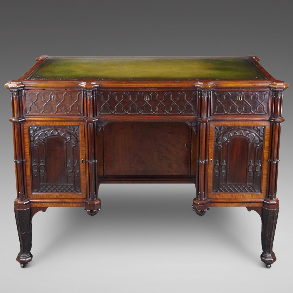 A Chippendale style dark mahogany Gothic Revival kneehole desk featuring carved tracery cupboard doors, a green leather writing surface, and carved legs.
