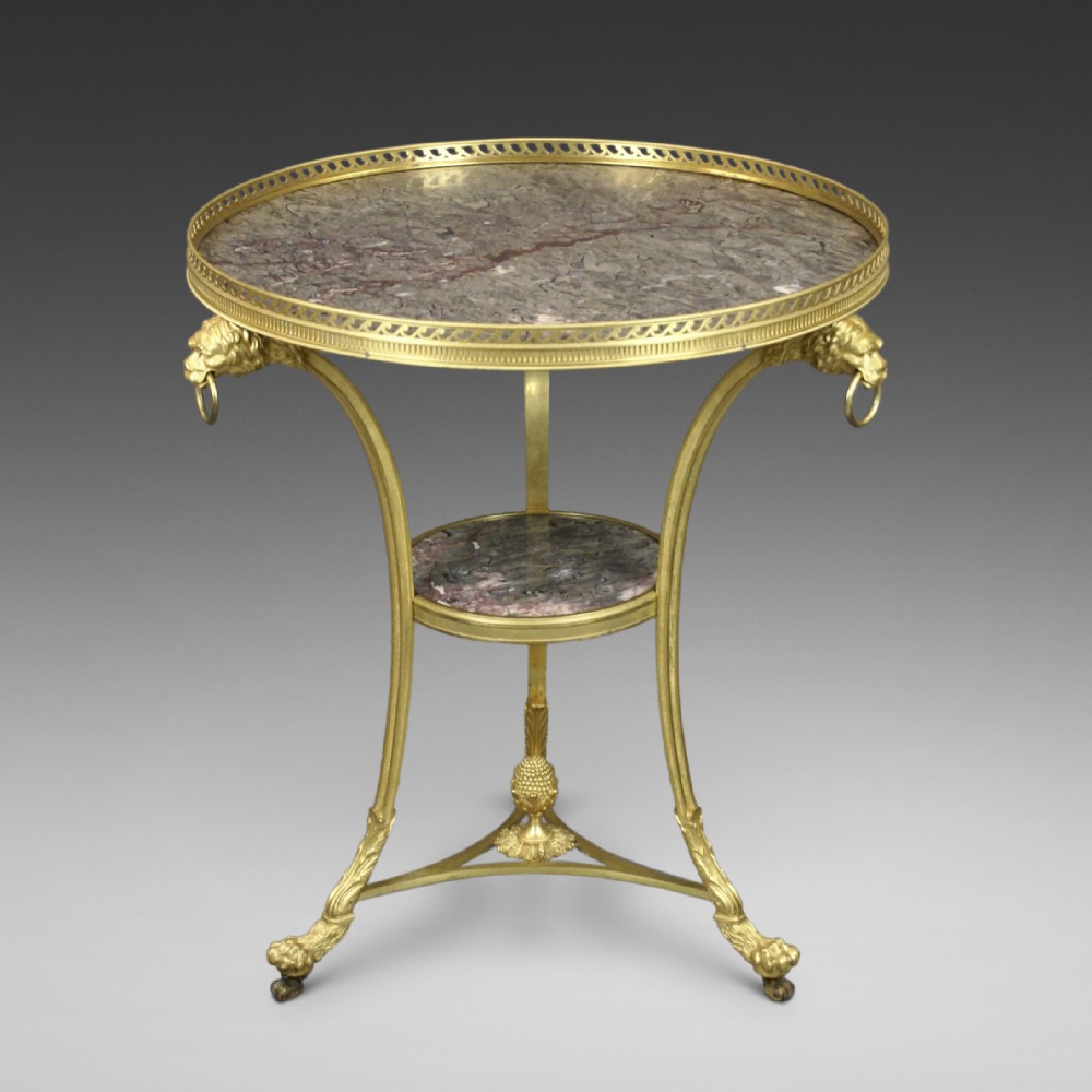 A circular French ormolu gueridon table featuring grey marble tops, a pierced gilt gallery, lion mask mounts with ring handles, and tripod legs terminating in paw feet.