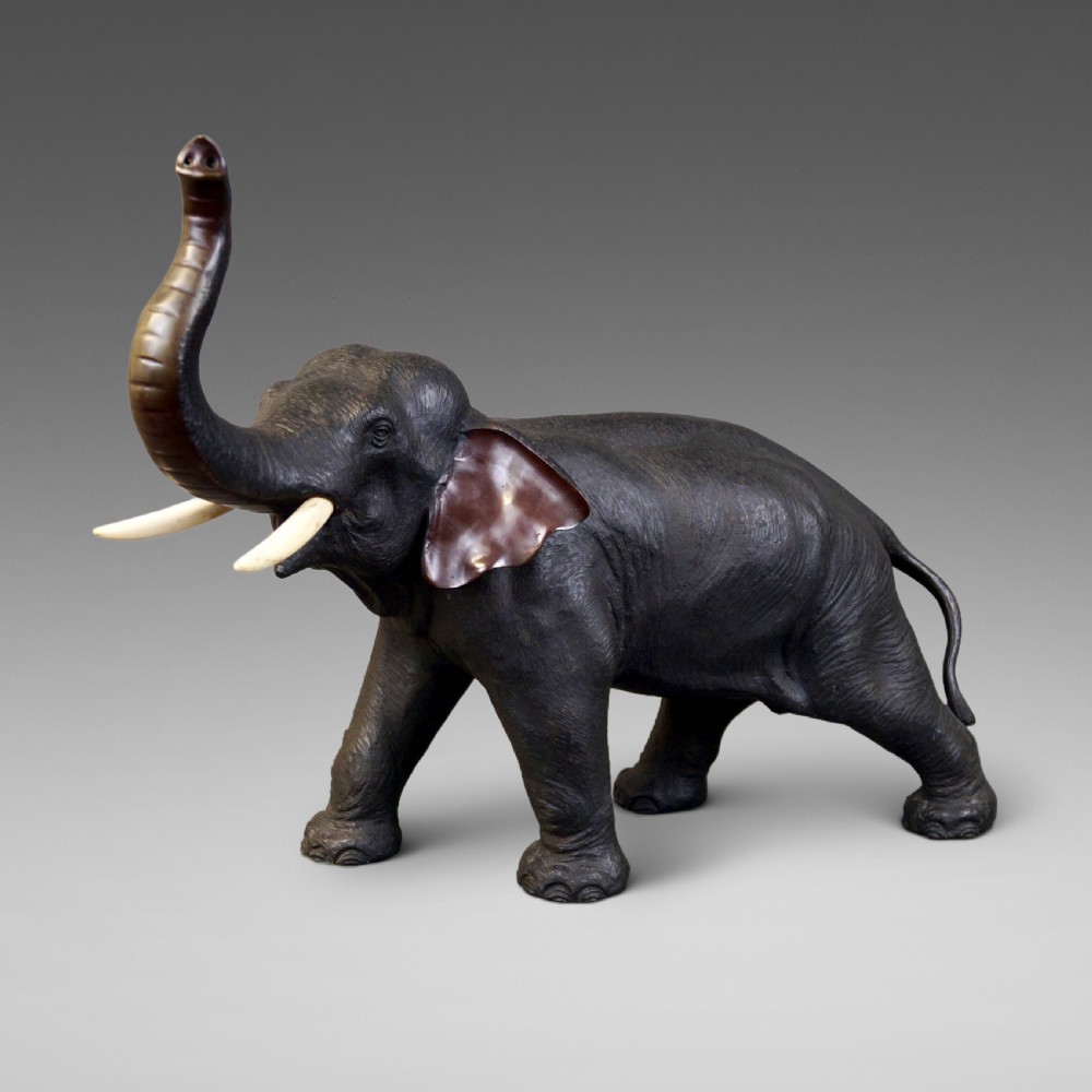 A dark brown cast bronze sculpture depicting an elephant with its trunk raised high and light-coloured tusks extended.