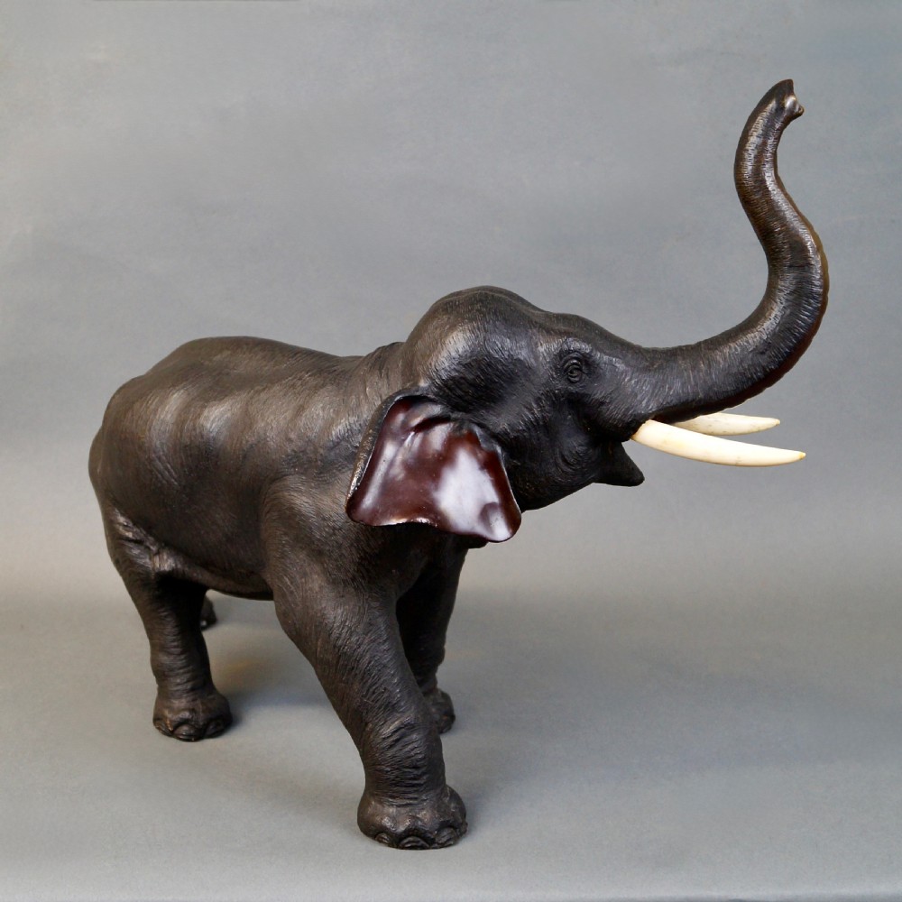 A dark brown cast bronze sculpture depicting an elephant with its trunk raised high and light-coloured tusks extended. View II