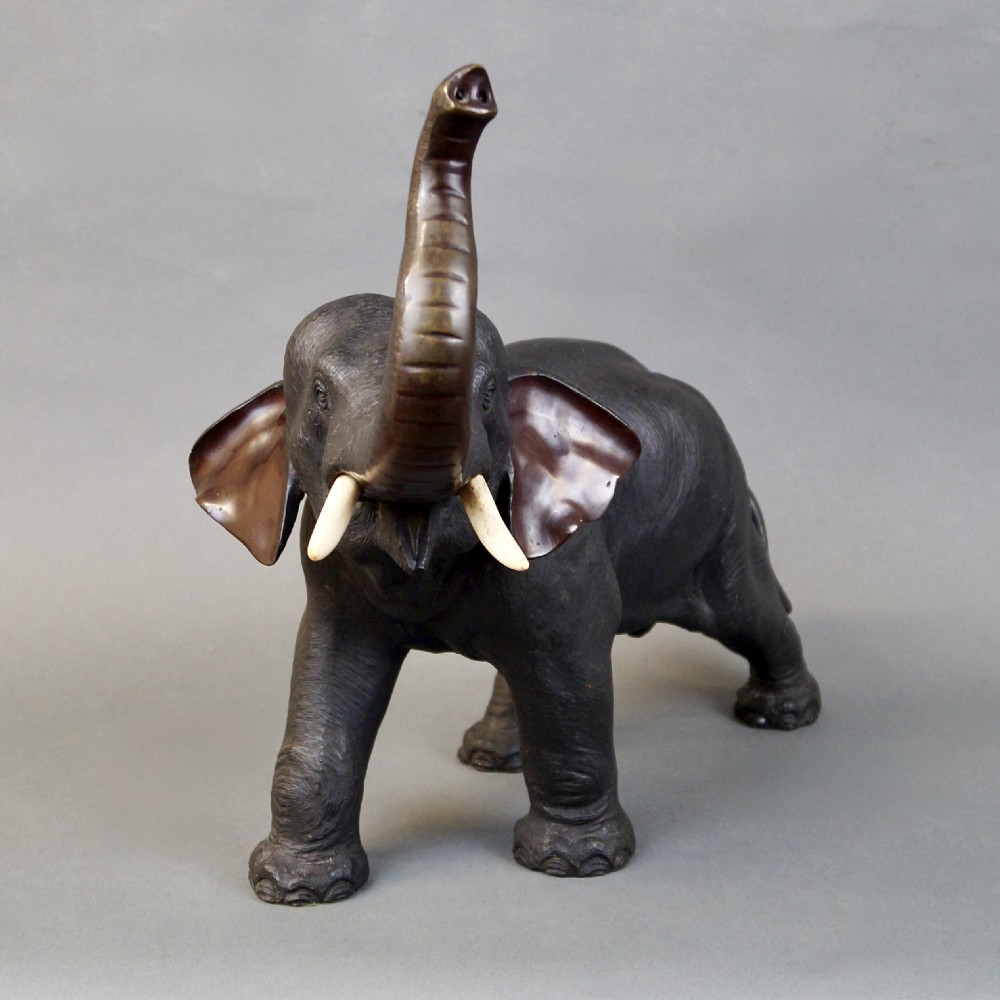 A dark brown cast bronze sculpture depicting an elephant with its trunk raised high and light-coloured tusks extended. View III