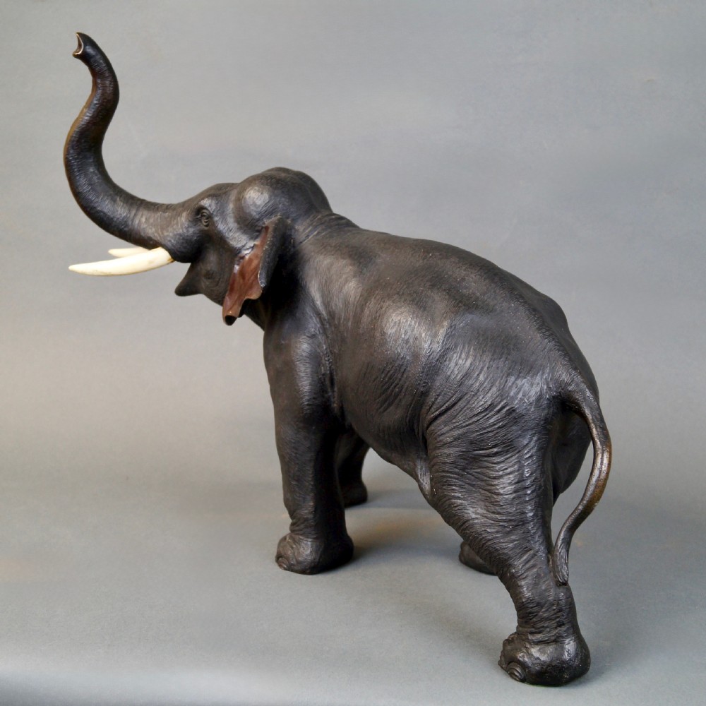 A dark brown cast bronze sculpture depicting an elephant with its trunk raised high and light-coloured tusks extended. View IV