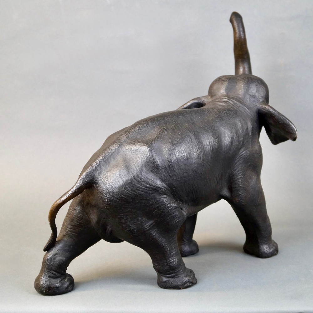 A dark brown cast bronze sculpture depicting an elephant with its trunk raised high and light-coloured tusks extended. View V
