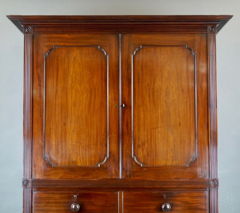 A Regency Cuban mahogany linen press featuring a two-door upper cupboard with panelled doors flanked by reeded pilasters over a lower chest of drawers raised on turned feet. View II