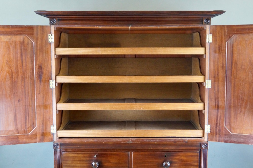 A Regency Cuban mahogany linen press featuring a two-door upper cupboard with panelled doors flanked by reeded pilasters over a lower chest of drawers raised on turned feet. View V