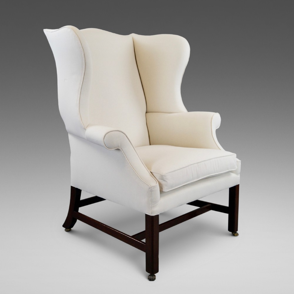 A George III mahogany wingback armchair upholstered in cream fabric, featuring straight dark mahogany legs joined by stretchers and set on brass castors.