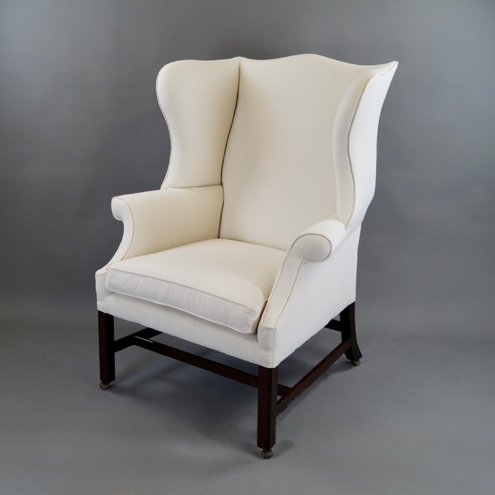 A George III mahogany wingback armchair upholstered in cream fabric, featuring straight dark mahogany legs joined by stretchers and set on brass castors. View II