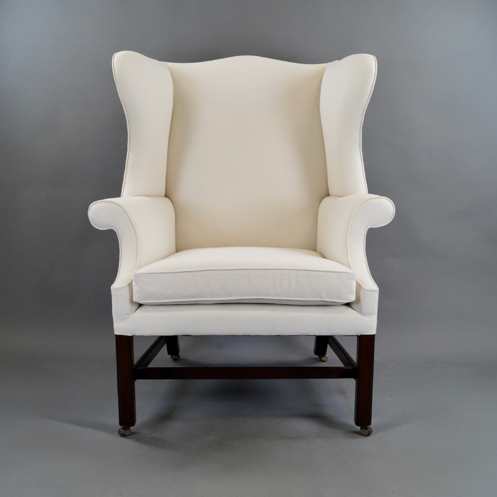 A George III mahogany wingback armchair upholstered in cream fabric, featuring straight dark mahogany legs joined by stretchers and set on brass castors. View III