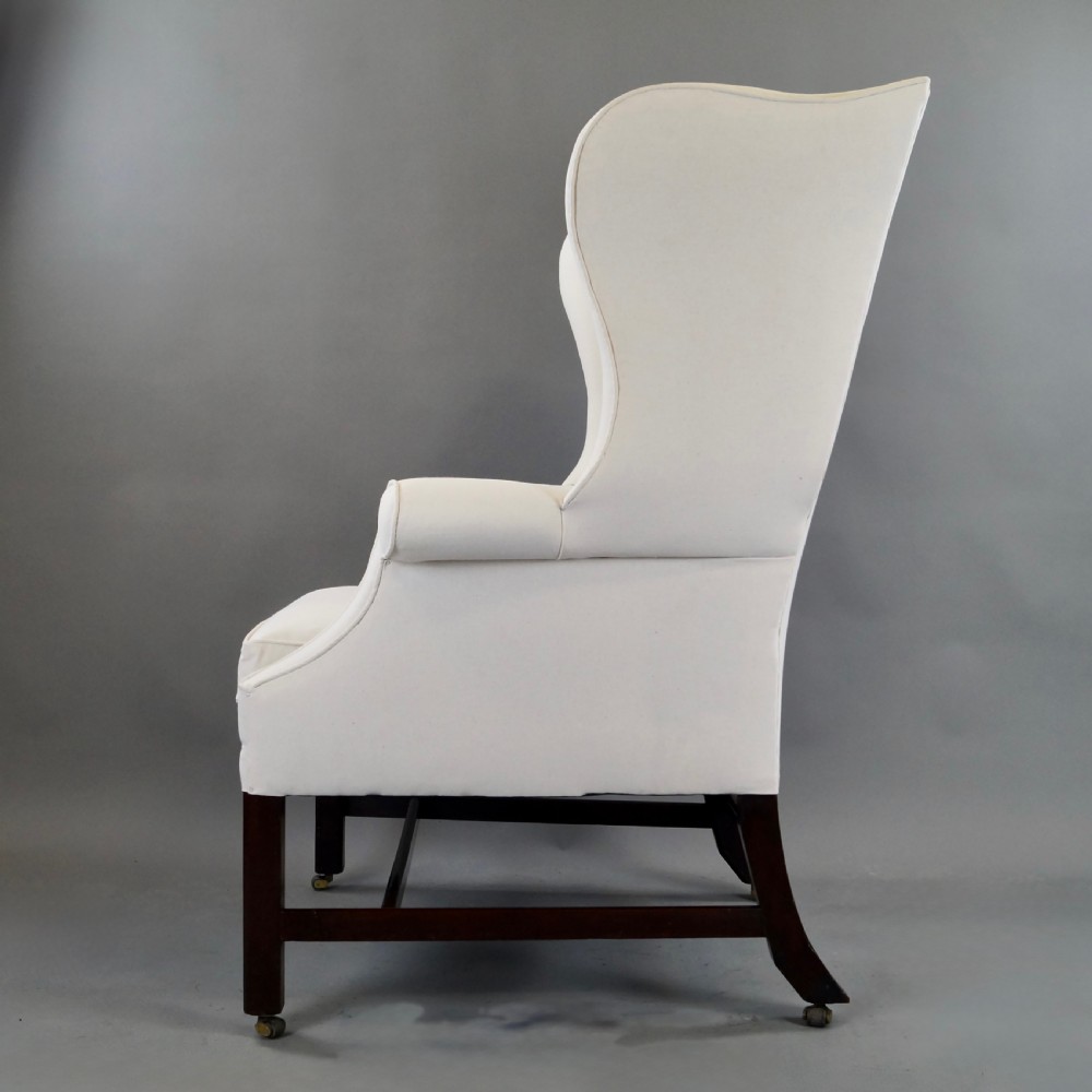 A George III mahogany wingback armchair upholstered in cream fabric, featuring straight dark mahogany legs joined by stretchers and set on brass castors. View V