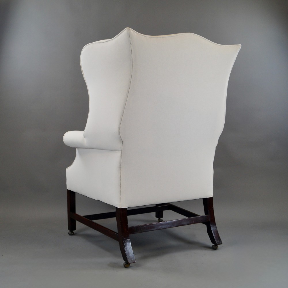 A George III mahogany wingback armchair upholstered in cream fabric, featuring straight dark mahogany legs joined by stretchers and set on brass castors. View VI