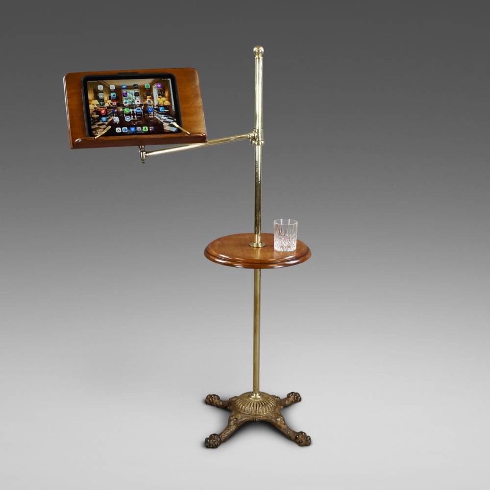 A late 19th-century Victorian reading stand with an adjustable brass stem, articulated arm holding a tilted mahogany book rest, a small central round mahogany shelf, and an ornate cast base.