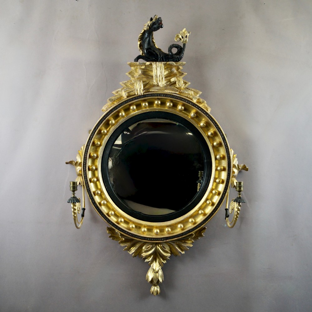 A circular Regency convex giltwood mirror with spherule embellishments, an ebonised dragon cresting, acanthus leaf apron, and twin candle sconces. View II