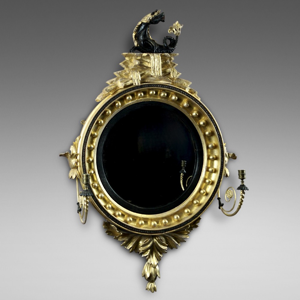 A circular Regency convex giltwood mirror with spherule embellishments, an ebonised dragon cresting, acanthus leaf apron, and twin candle sconces.