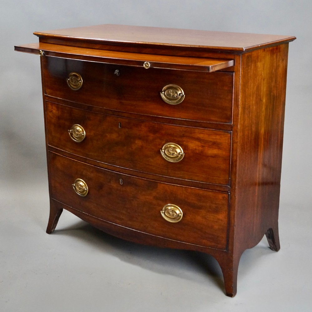 A George III mahogany bow-fronted chest of drawers featuring three graduated drawers with brass oval handles, a upper brushing slide, and splayed bracket feet. View II
