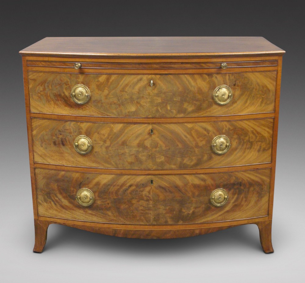 A George III bow-fronted mahogany chest of drawers with satinwood crossbanding, three long drawers, flame veneer panels, and brass ring handles.