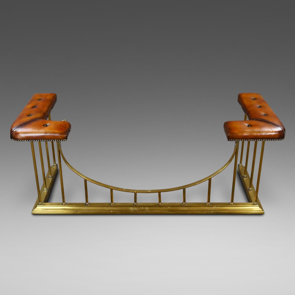 A nineteenth-century brass club fender featuring a central dipped rail, vertical brass spindles, and button-tufted tan leather corner seats.