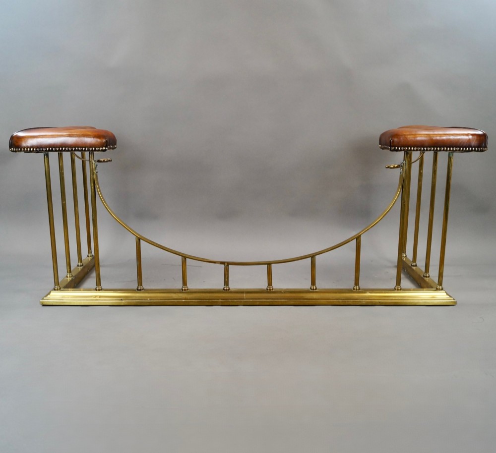 A nineteenth-century brass club fender featuring a central dipped rail, vertical brass spindles, and button-tufted tan leather corner seats. View V