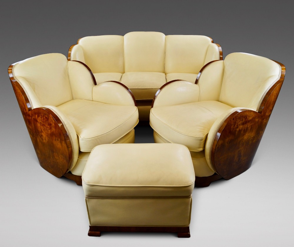 An Art Deco cloud-back lounge suite comprising a three-seater sofa, two matching armchairs, and an ottoman, all upholstered in cream leather with dark burr walnut bentwood outer panels.