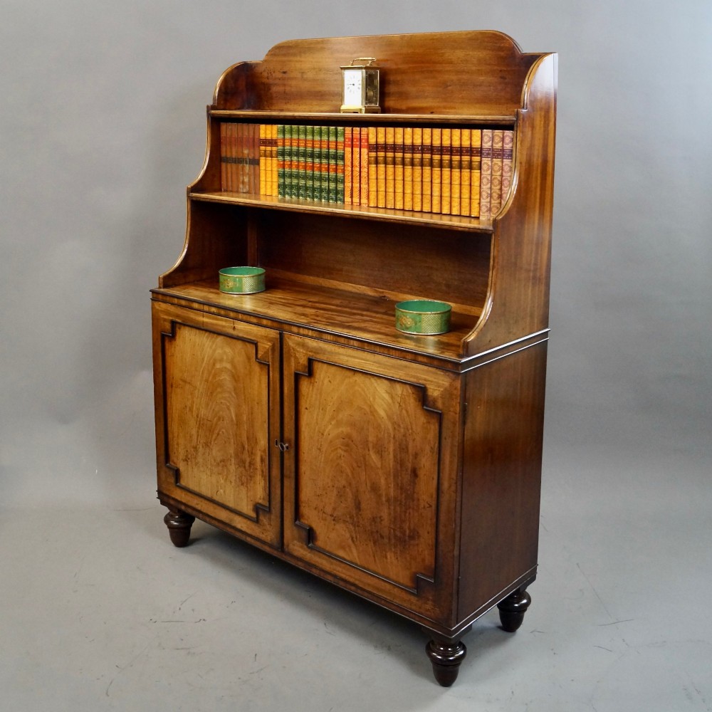 An antique Regency mahogany waterfall bookcase featuring open graduated shelves above a two-door lower cupboard resting on turned legs. View II