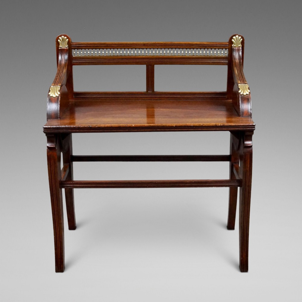 A dark mahogany bench with a brass spindle gallery rail, brass corner mounts, and straight legs joined by stretchers.