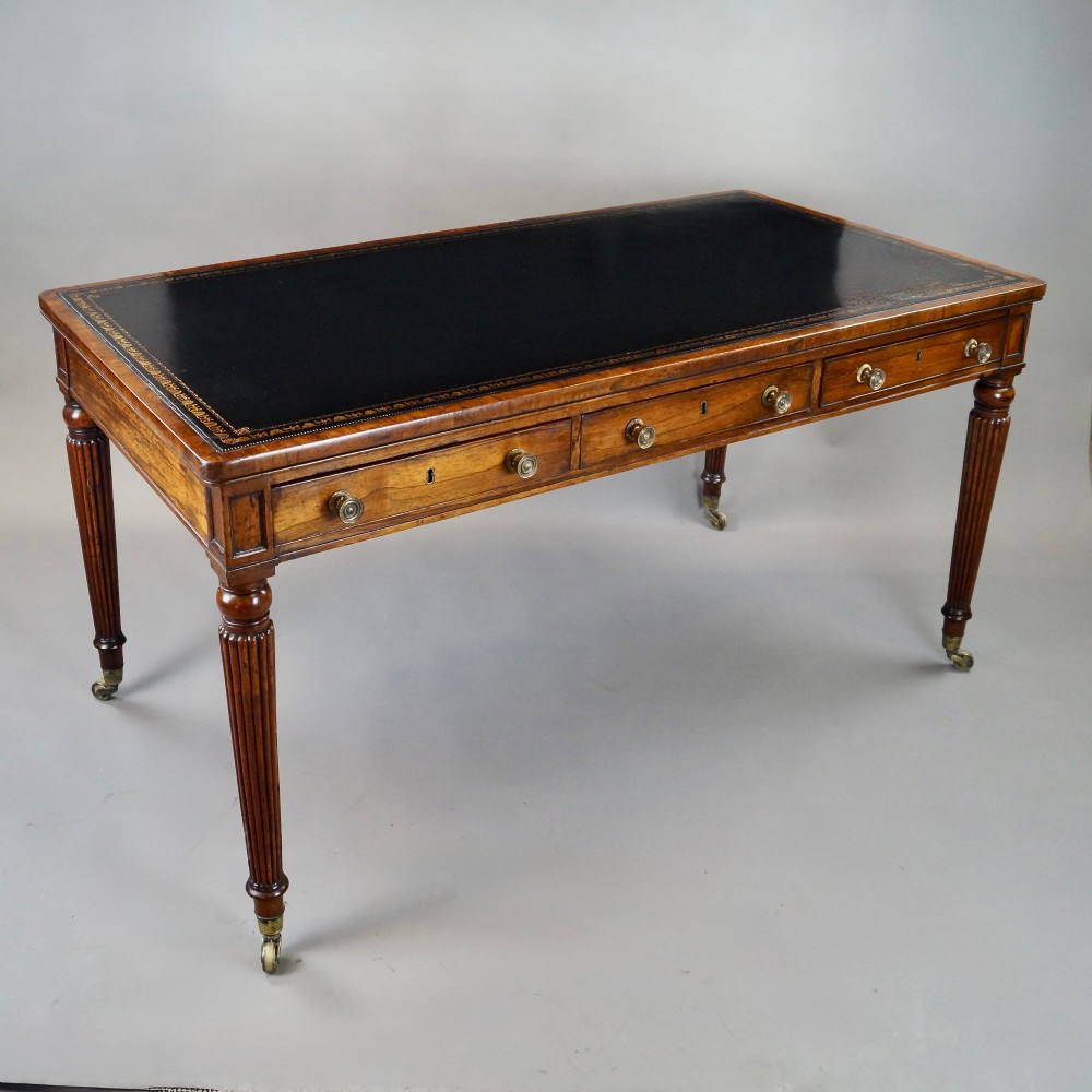 A Regency rosewood writing table featuring a dark leather inset top with gold tooling, three frieze drawers with brass knobs, and reeded legs on brass castors.