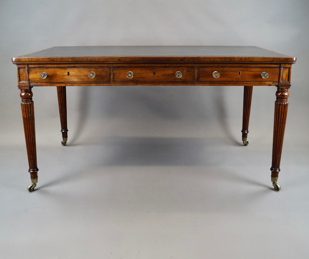 A Regency rosewood writing table featuring a dark leather inset top with gold tooling, three frieze drawers with brass knobs, and reeded legs on brass castors. View II