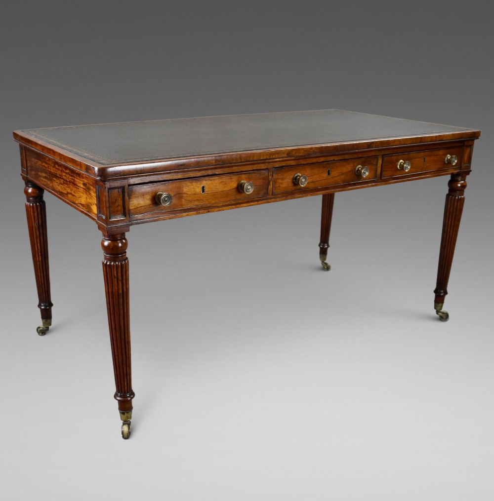 A Regency rosewood writing table featuring a dark leather inset top with gold tooling, three frieze drawers with brass knobs, and reeded legs on brass castors. View III