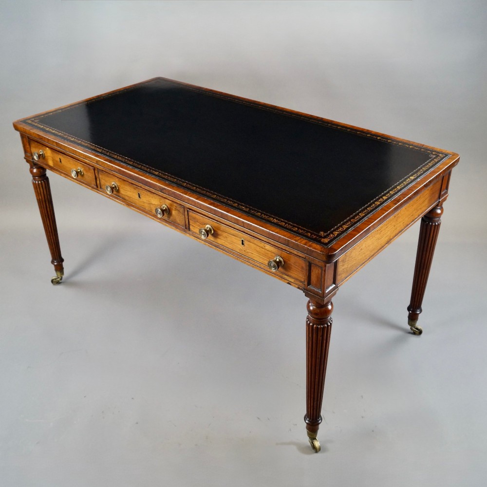 A Regency rosewood writing table featuring a dark leather inset top with gold tooling, three frieze drawers with brass knobs, and reeded legs on brass castors. View IV