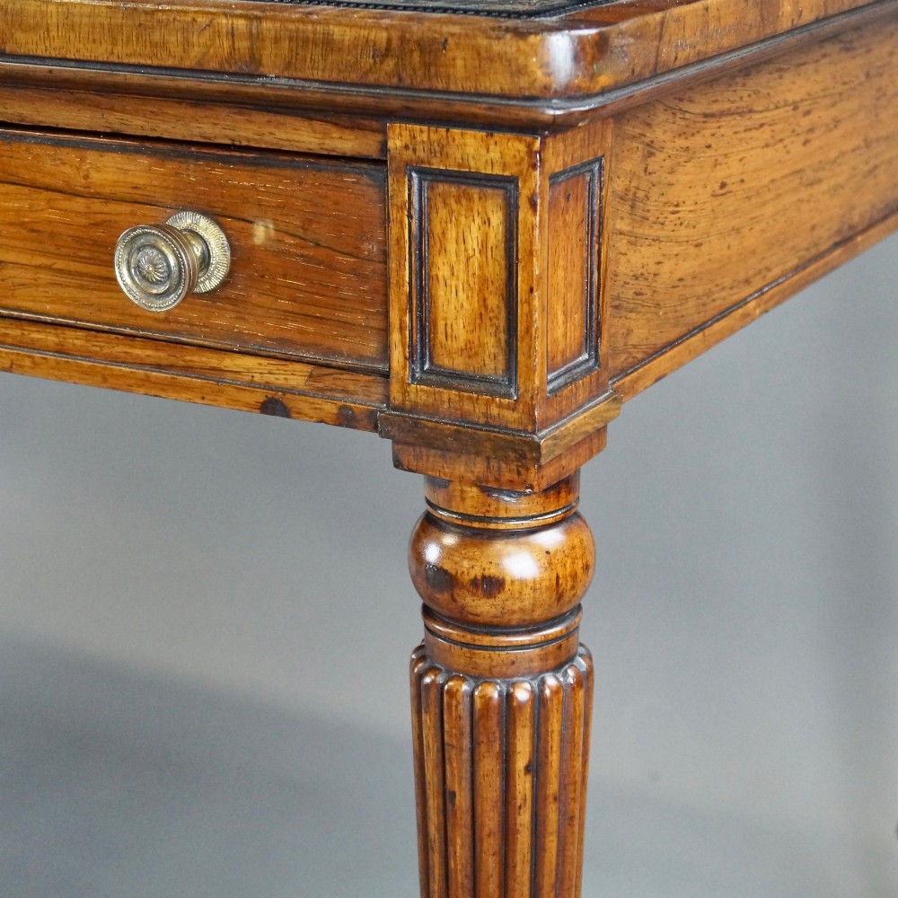 A Regency rosewood writing table featuring a dark leather inset top with gold tooling, three frieze drawers with brass knobs, and reeded legs on brass castors. View V