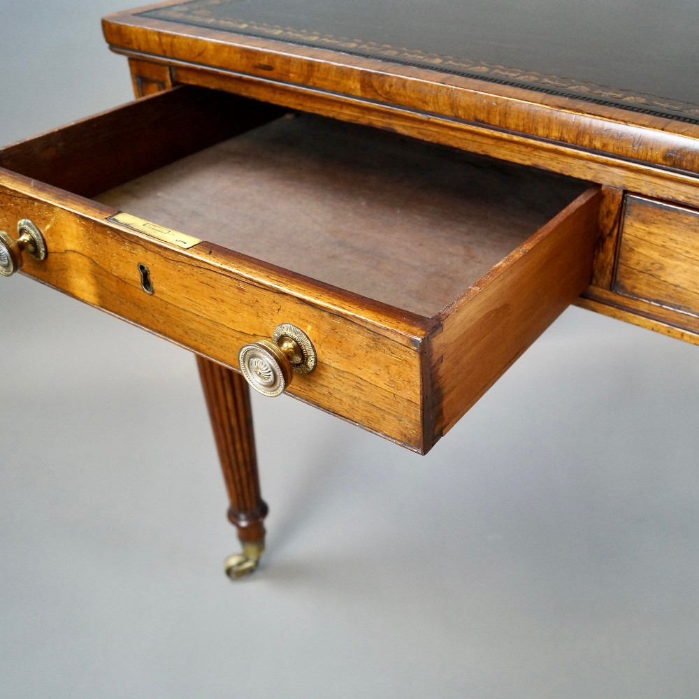 A Regency rosewood writing table featuring a dark leather inset top with gold tooling, three frieze drawers with brass knobs, and reeded legs on brass castors. View VI