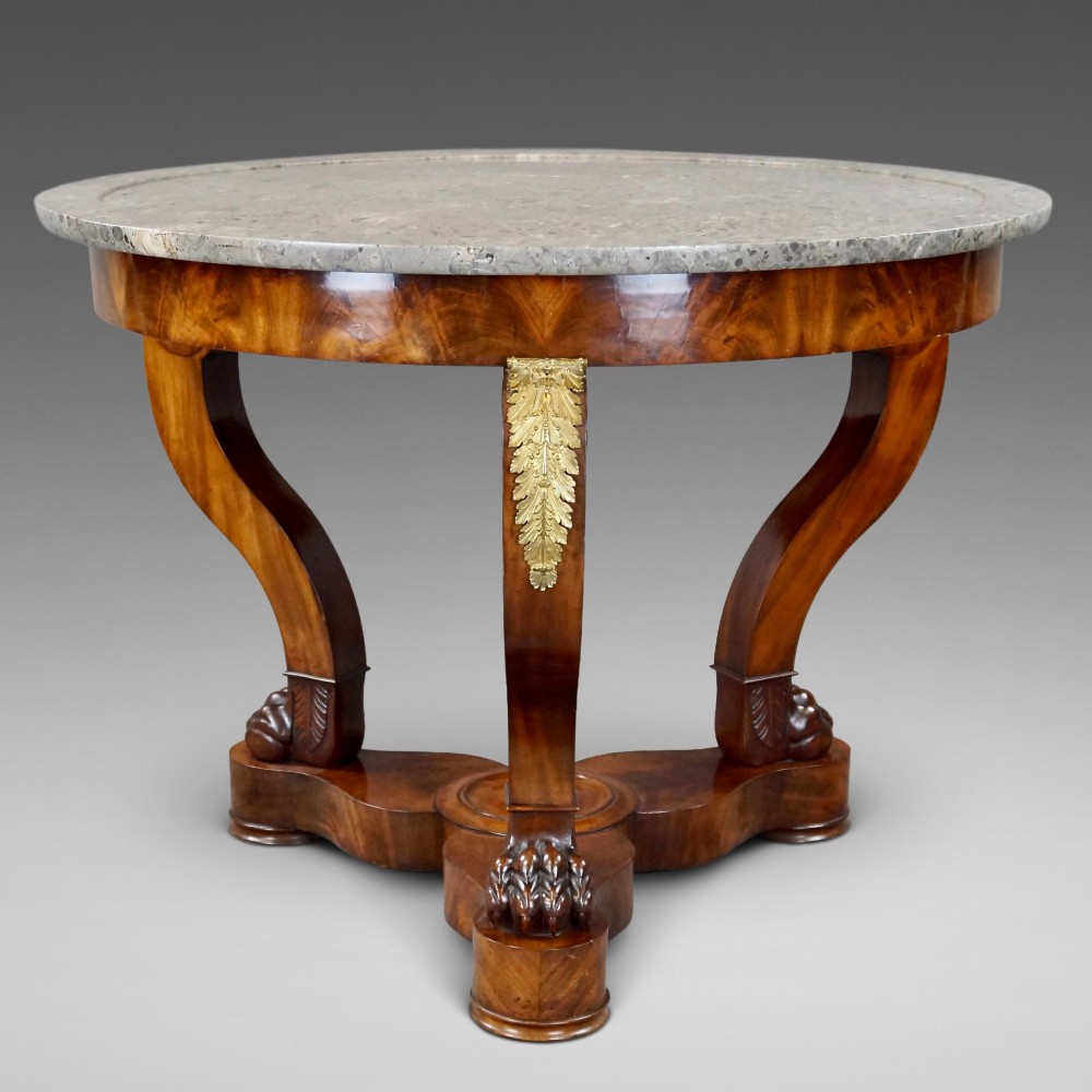 A circular French Empire mahogany gueridon table featuring a grey marble top, gilt bronze mounts, curved tripod legs, and carved lion paw feet on a plinth base.