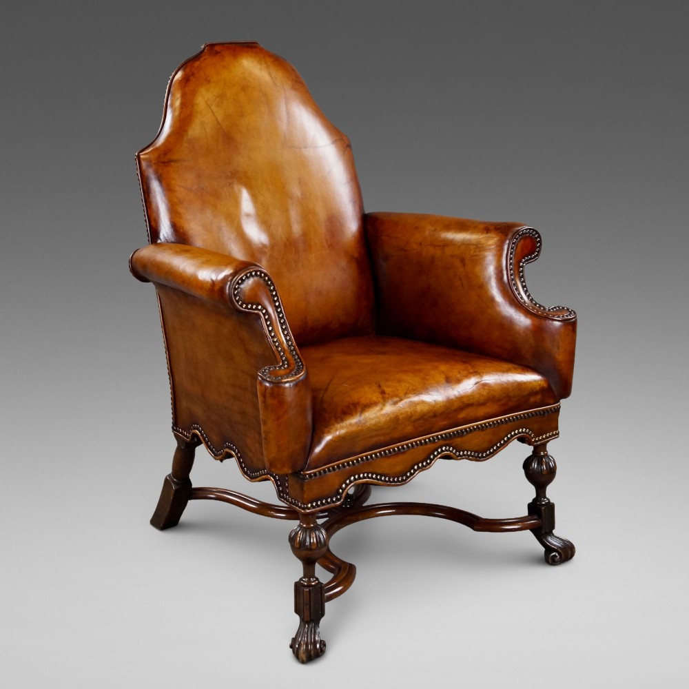 An 18th-century style tan leather armchair with brass stud trimming, scrolled arms, a shaped backrest, and carved walnut legs with a curved stretcher.