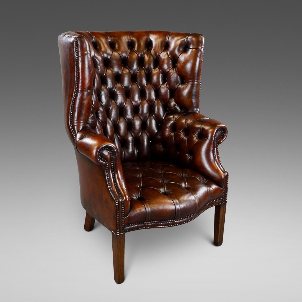 A chestnut brown leather wingback armchair featuring deep button tufting, brass stud detailing along the borders, and straight wooden legs.