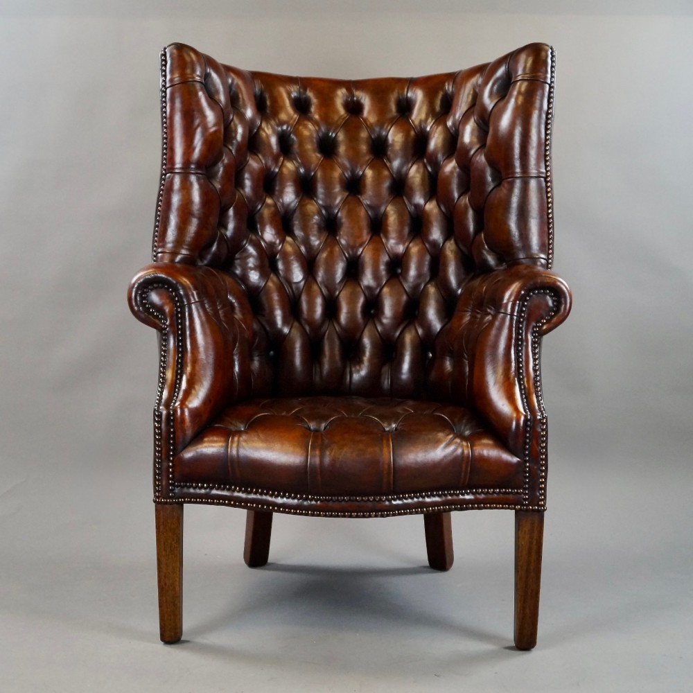 A chestnut brown leather wingback armchair featuring deep button tufting, brass stud detailing along the borders, and straight wooden legs. View III