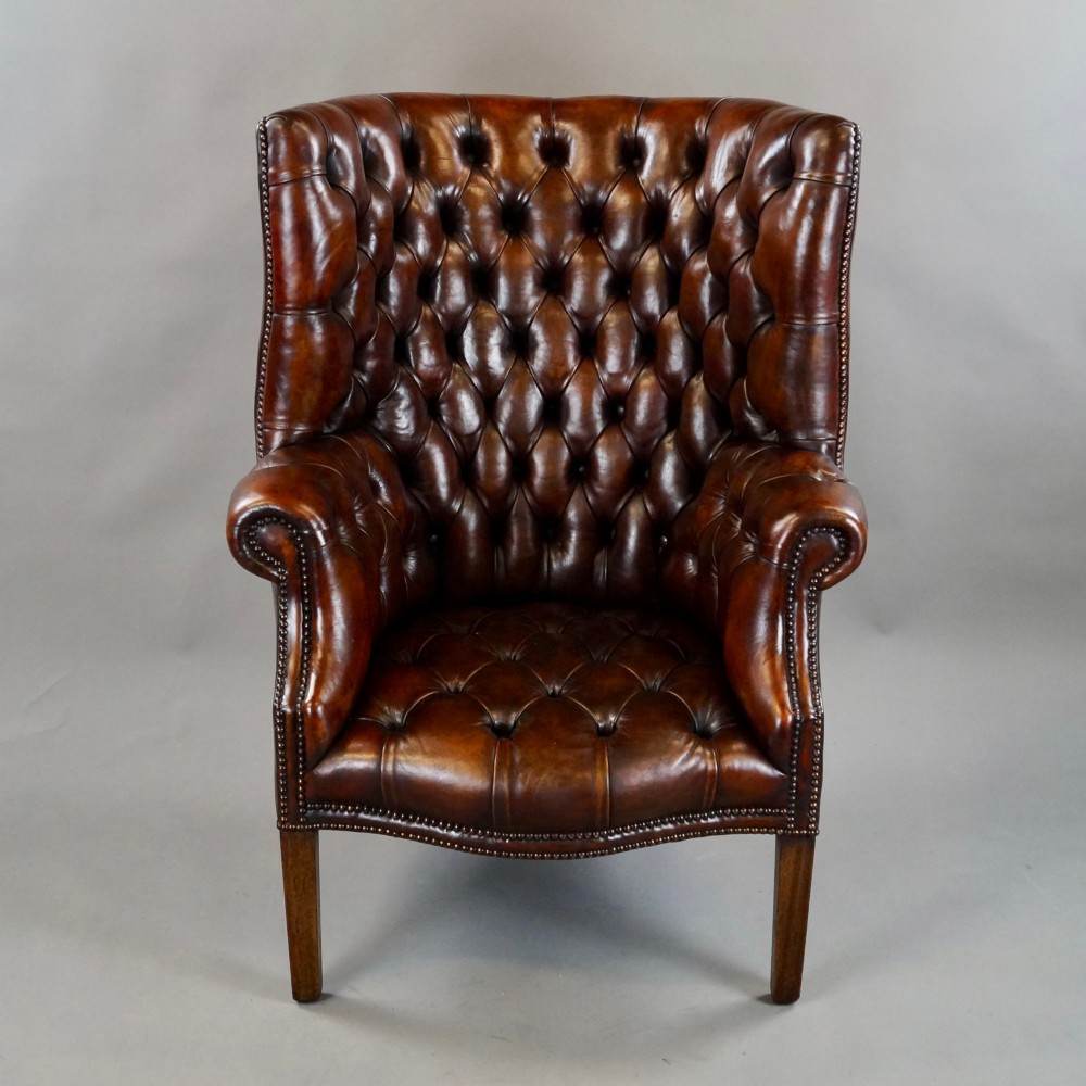 A chestnut brown leather wingback armchair featuring deep button tufting, brass stud detailing along the borders, and straight wooden legs. View V