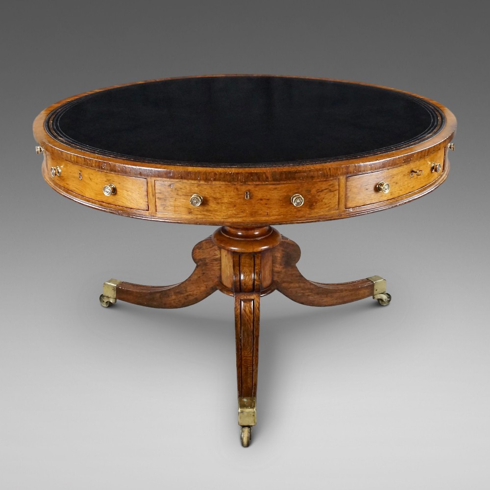 A circular oak library drum table featuring a black leather surface insert, frieze drawers with small round brass knobs, and a central pedestal base with four splayed legs on brass castors.