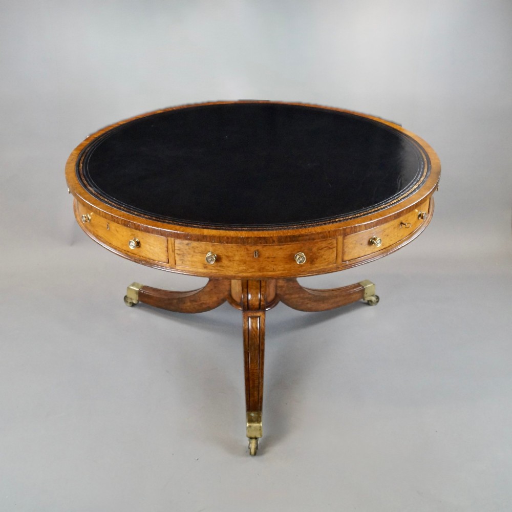 A circular oak library drum table featuring a black leather surface insert, frieze drawers with small round brass knobs, and a central pedestal base with four splayed legs on brass castors. View II