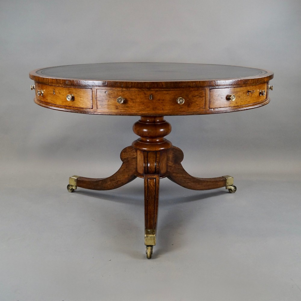 A circular oak library drum table featuring a black leather surface insert, frieze drawers with small round brass knobs, and a central pedestal base with four splayed legs on brass castors. View III