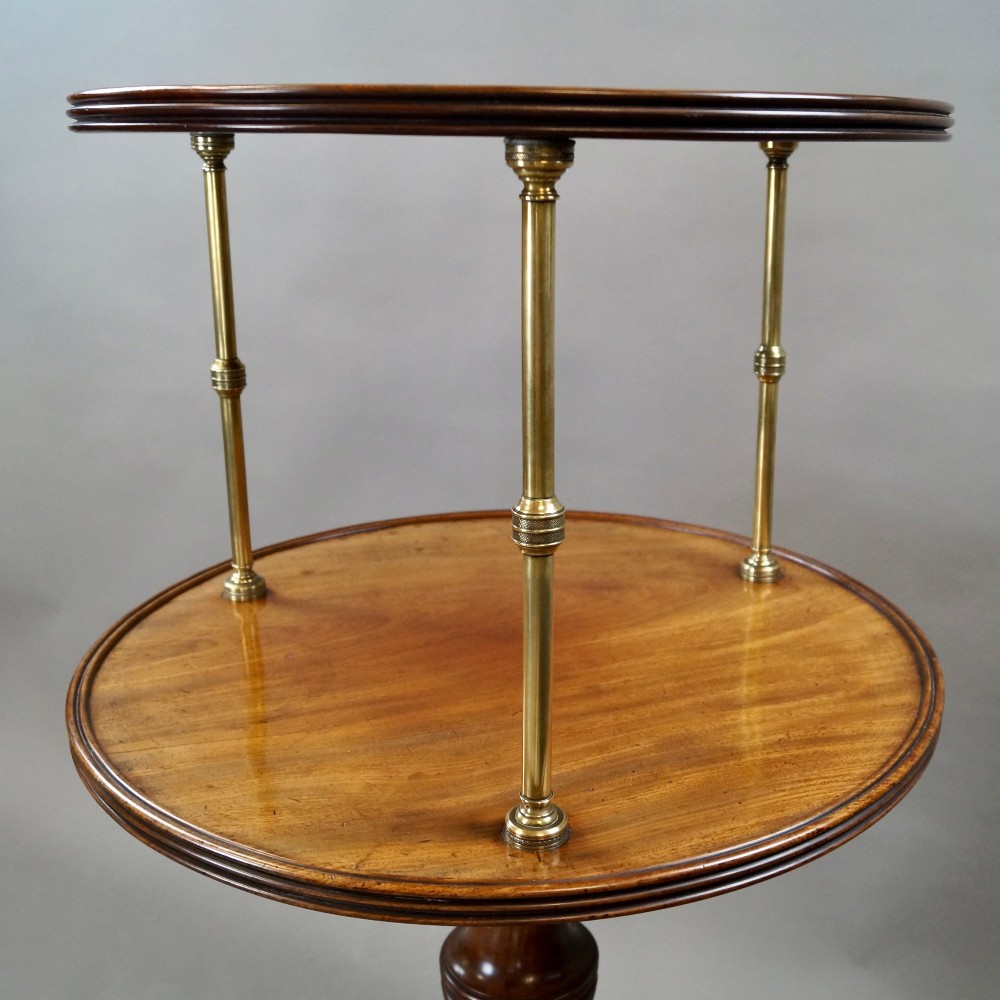 A two-tier Regency mahogany dumb waiter featuring circular surfaces separated by turned brass columns and supported on a turned central pedestal with tripod legs and brass castors. View V