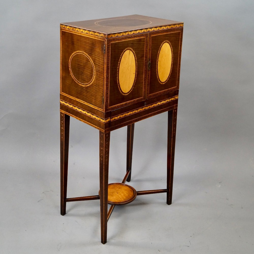 A Sheraton period harewood cabinet on stand featuring twin cupboard doors with oval satinwood inlays and slender tapered legs connected by a circular stretcher tray. View III