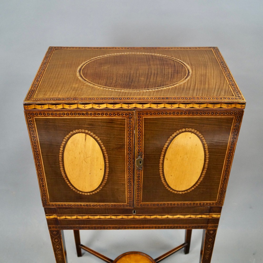 A Sheraton period harewood cabinet on stand featuring twin cupboard doors with oval satinwood inlays and slender tapered legs connected by a circular stretcher tray. View V